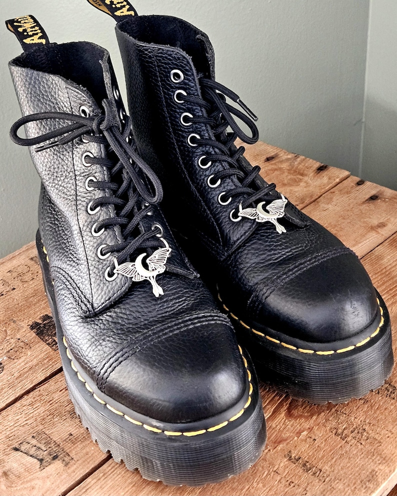 May include: A pair of black leather platform boots with silver-tone moth charms on the laces. The boots have a textured finish, yellow stitching, and black laces. The boots are on a wooden surface.