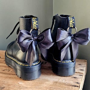 May include: Pair of black leather platform boots with black satin bows. The boots feature yellow stitching and the words "AirWair" in yellow on the pull tab. The boots are on a wooden surface.