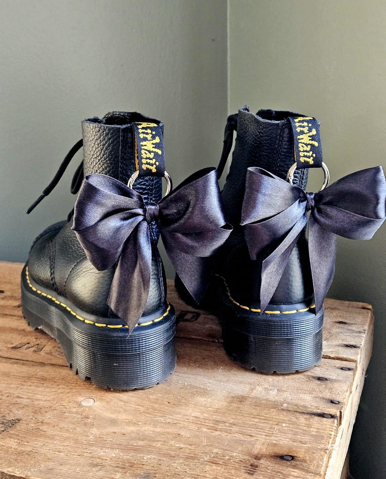 May include: Pair of black leather platform boots with black satin bows. The boots feature yellow stitching and the words "AirWair" in yellow on the pull tab. The boots are on a wooden surface.