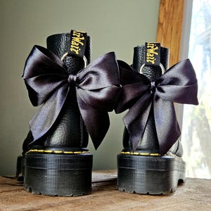 May include: Two black platform boots with large black satin bows. The boots feature yellow stitching and "Air Wair" in gold on the pull tab. The boots are displayed on a wooden surface.
