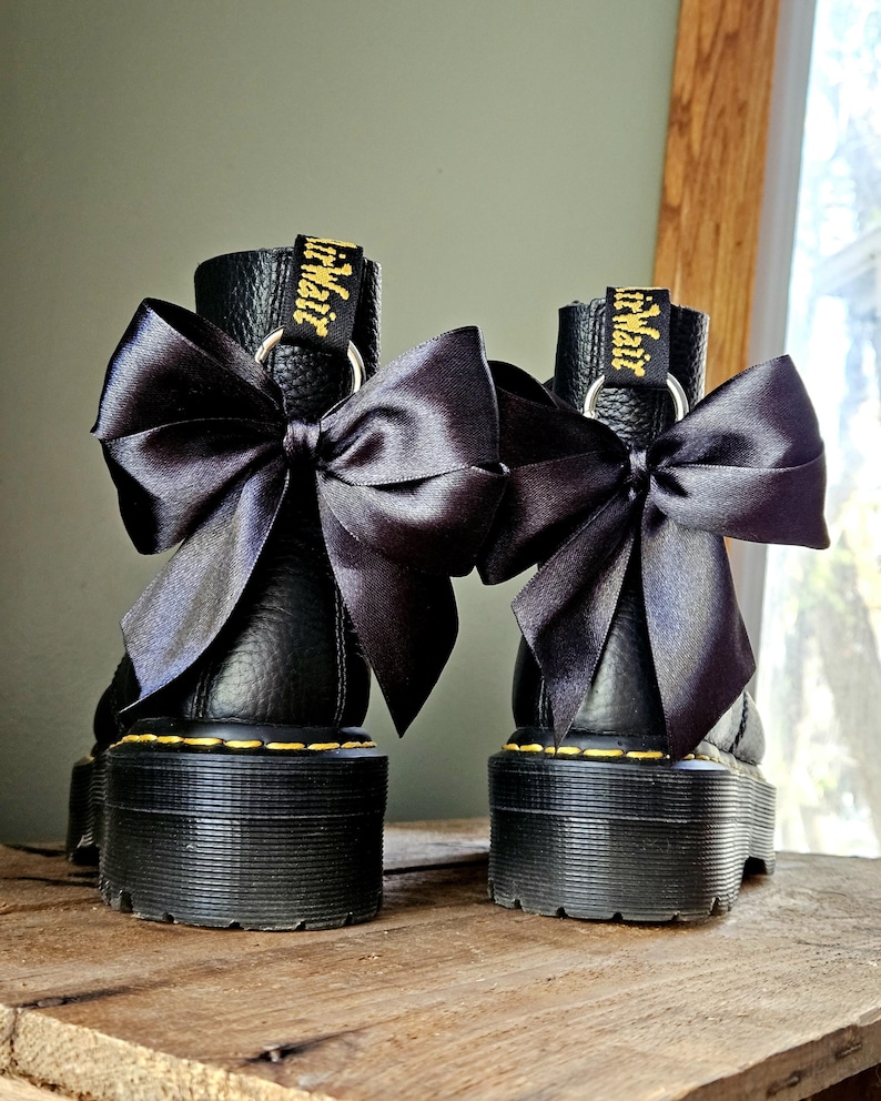 May include: Two black platform boots with large black satin bows. The boots feature yellow stitching and "Air Wair" in gold on the pull tab. The boots are displayed on a wooden surface.