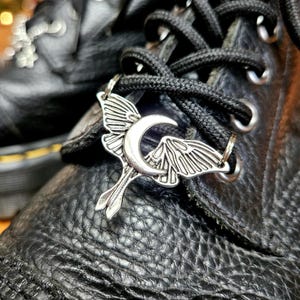 May include: Close-up of black leather boots with black laces. A silver-colored charm in the shape of a moth with a crescent moon is attached to the laces. The boots have a textured surface.