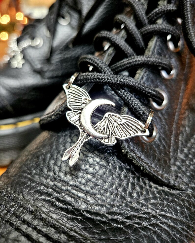 May include: Close-up of black leather boots with black laces. A silver-colored charm in the shape of a moth with a crescent moon is attached to the laces. The boots have a textured surface.