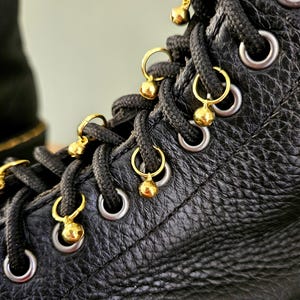 May include: Close-up of a black leather boot with black laces and silver eyelets. Gold-colored charms with small spheres hang from the laces, adding a decorative touch. The leather has a textured appearance.