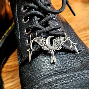 May include: Close-up of a black leather boot with black laces. Silver charms hang from the laces, including a moth with a crescent moon, a star, and a four-pointed star. The boot has visible stitching.