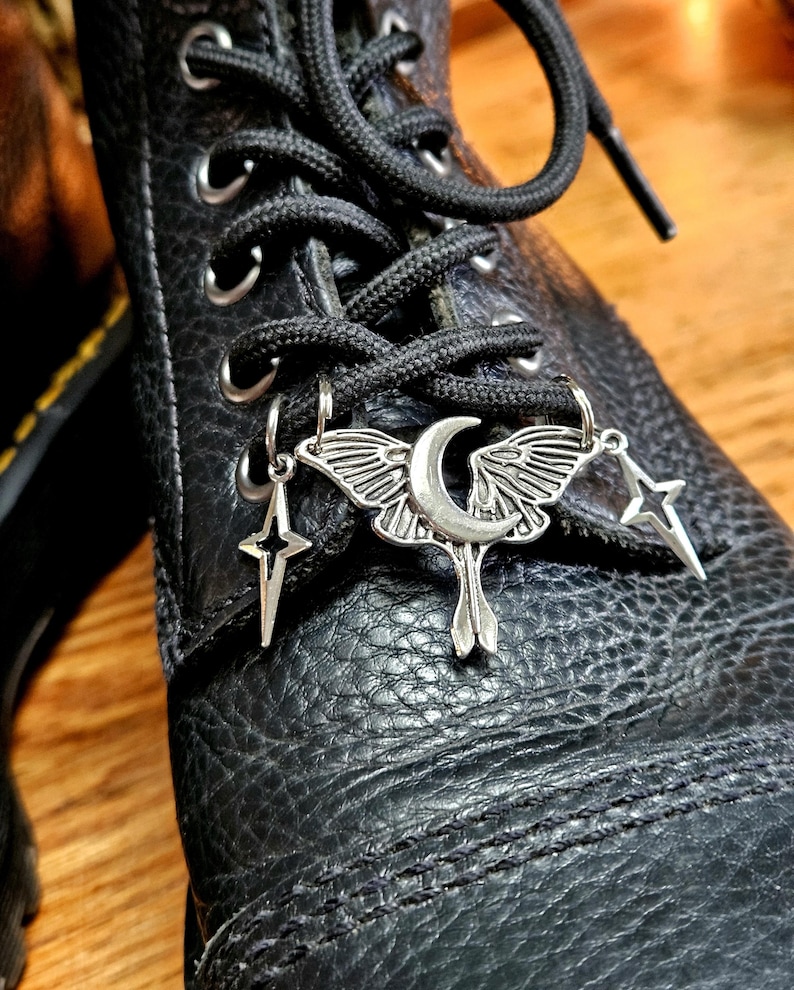 May include: Close-up of a black leather boot with black laces. Silver charms hang from the laces, including a moth with a crescent moon, a star, and a four-pointed star. The boot has visible stitching.