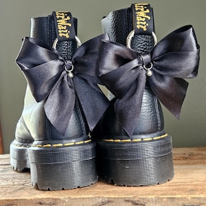 Puede incluir: Botas de plataforma de cuero negro con lazos de satén negro. Las botas tienen costuras amarillas y la marca "AirWair" en la lengüeta. Las botas están sobre una superficie de madera. Miden unos 10 cm de alto.