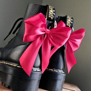 Może przedstawiać: Czarne buty na platformie z jasnoróżowymi satynowymi kokardami przymocowanymi do tylnych pętelek. Buty mają żółte szwy, a nazwa marki jest widoczna na uchwytach.