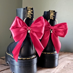 May include: Two black platform boots with vibrant red satin bows. The boots feature yellow stitching and gold lettering on the pull tabs. The boots are displayed on a wooden surface. The boots are approximately 10 cm high.
