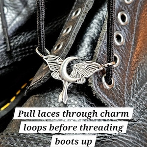 May include: A silver-colored boot charm in the shape of a crescent moon with wings, hanging from a black shoelace. The charm is placed on a black leather boot with visible stitching and eyelets. Text on the image reads: "Pull laces through charm loops before threading boots up."