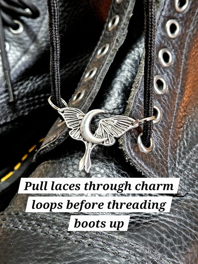 May include: A silver-colored boot charm in the shape of a crescent moon with wings, hanging from a black shoelace. The charm is placed on a black leather boot with visible stitching and eyelets. Text on the image reads: "Pull laces through charm loops before threading boots up."