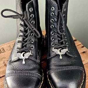 May include: A pair of black leather boots with silver-toned moth-shaped charms on the laces. The boots have a textured finish, black laces, and yellow stitching around the soles. The boots are on a wooden surface.