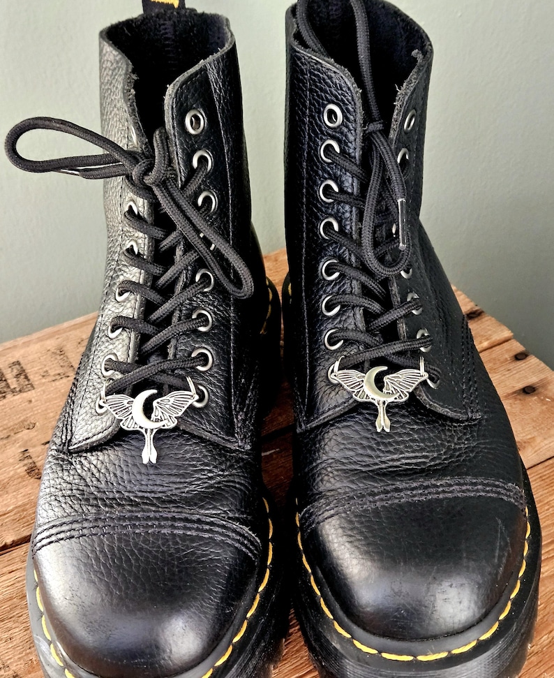 May include: A pair of black leather boots with silver-toned moth-shaped charms on the laces. The boots have a textured finish, black laces, and yellow stitching around the soles. The boots are on a wooden surface.