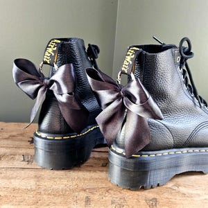 May include: Two black leather platform boots with dark grey satin bows. The boots feature yellow stitching and "AirWair" branding on the pull tabs. The boots are a stylish footwear choice.