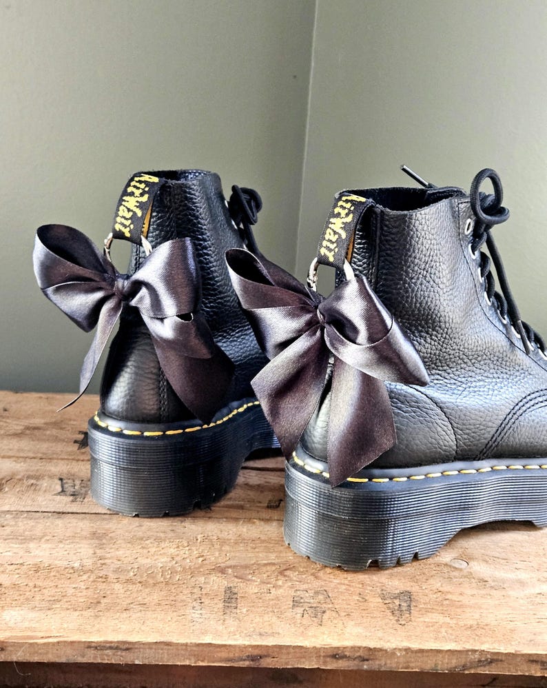 May include: Two black leather platform boots with dark grey satin bows. The boots feature yellow stitching and "AirWair" branding on the pull tabs. The boots are a stylish footwear choice.