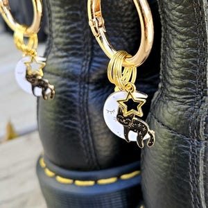 Könnte beinhalten: Nahaufnahme von schwarzen Lederstiefeln mit goldfarbener Hardware. Jeder Stiefel hat einen goldenen Anhänger mit einem weißen Mond, einer schwarzen Katze und einem goldenen Stern. Die Stiefel haben gelbe Nähte.
