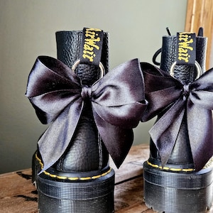 May include: A pair of black leather boots with chunky soles, each decorated with a large black satin bow. The boots feature yellow stitching and a yellow and black tag reading "AirWair". They are set on a wooden surface.
