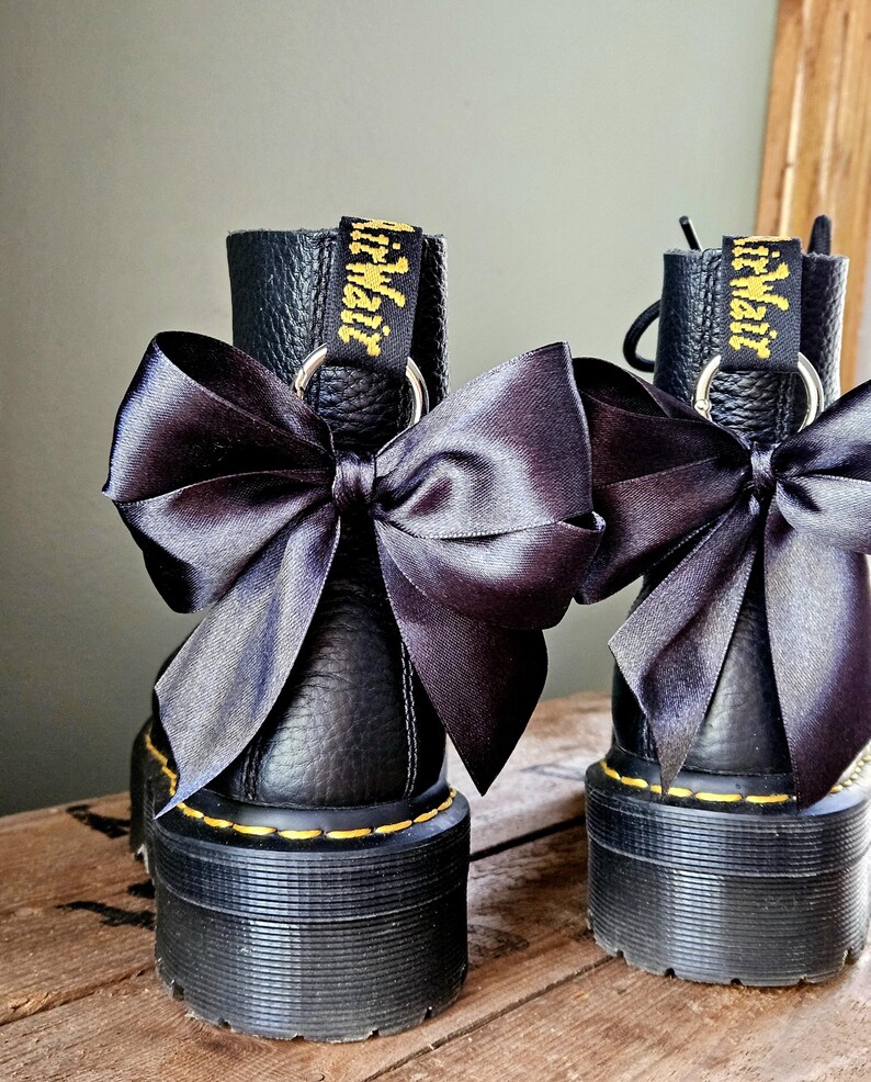 May include: A pair of black leather boots with chunky soles, each decorated with a large black satin bow. The boots feature yellow stitching and a yellow and black tag reading "AirWair". They are set on a wooden surface.