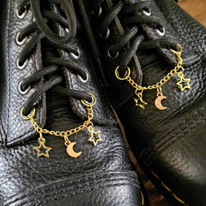 May include: Black leather boots with gold-colored star and crescent moon charms hanging from the laces. The boots have black laces and yellow stitching along the soles. The charms are connected by a delicate chain.