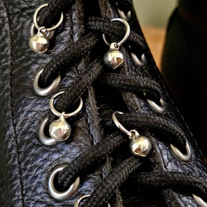 Pode incluir: Uma bota de couro preto texturizado com atacadores pretos enfiados através de ilhós de metal prateado. Pequenos guizos redondos de metal prateado estão presos aos atacadores usando anéis de metal circulares em cada ilhó.
