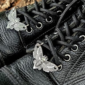 Pode incluir: Um par de botas de couro preto com adornos metálicos prateados em forma de traça para os atacadores. Cada adorno apresenta padrões gravados intrincados nas asas e um pequeno detalhe de caveira no centro, preso aos atacadores pretos com anéis.