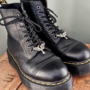 May include: Pair of black leather platform boots with black laces and silver-tone charms. The boots have yellow stitching around the soles and the brand name "Airwair" is visible on the pull tabs. The boots are on a wooden surface.