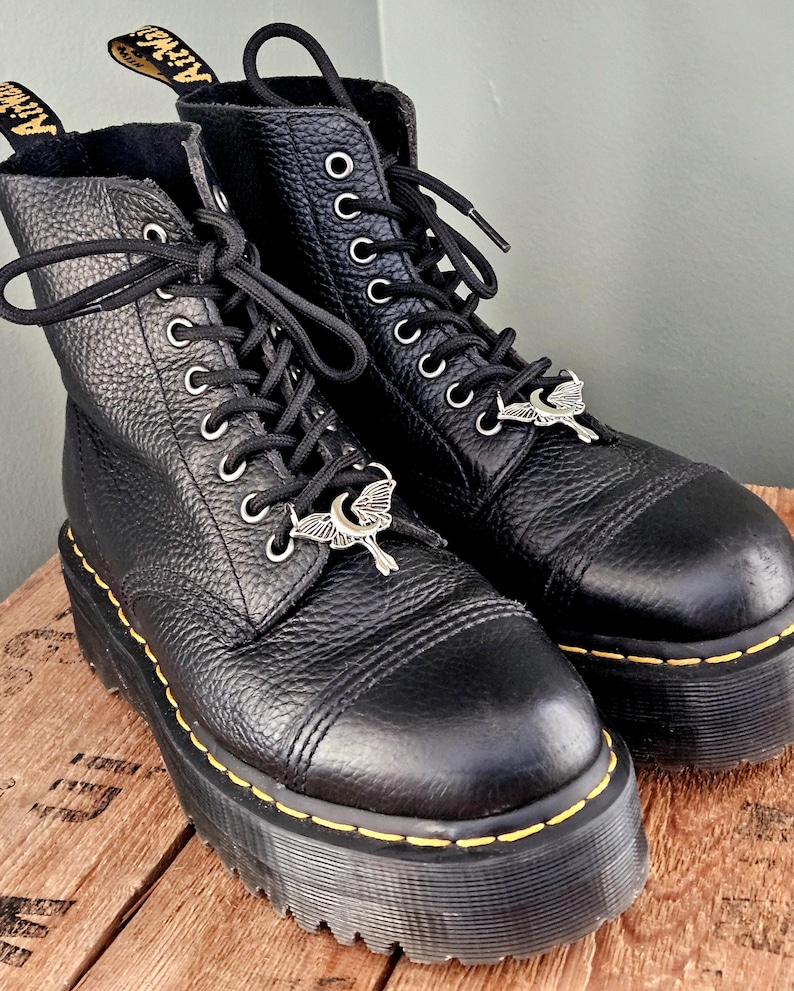 May include: Pair of black leather platform boots with black laces and silver-tone charms. The boots have yellow stitching around the soles and the brand name "Airwair" is visible on the pull tabs. The boots are on a wooden surface.