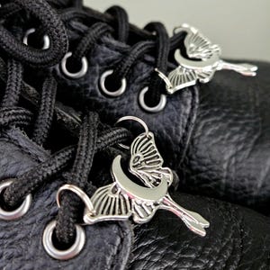 May include: Close-up of black leather boots with black laces and silver-colored butterfly-shaped charms. The charms are attached to the laces, adding a decorative element to the footwear. The boots have metal eyelets and a textured leather surface.