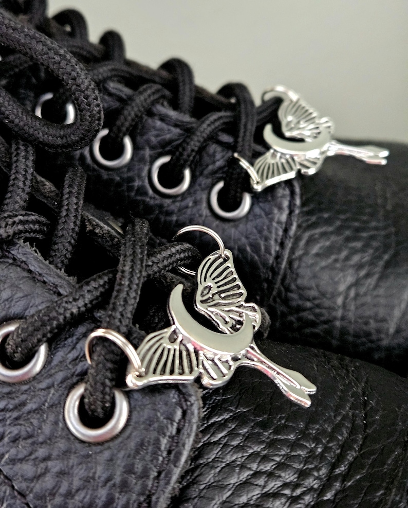 May include: Close-up of black leather boots with black laces and silver-colored butterfly-shaped charms. The charms are attached to the laces, adding a decorative element to the footwear. The boots have metal eyelets and a textured leather surface.