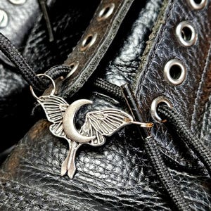 May include: Close-up of a silver-colored moth and crescent moon charm on a black boot. The charm is attached to the black shoelaces. The boot is made of textured black leather with metal eyelets.