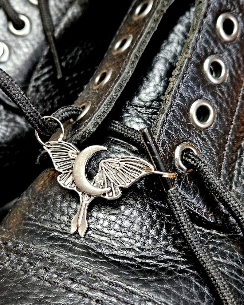 May include: Close-up of a silver-colored moth and crescent moon charm on a black boot. The charm is attached to the black shoelaces. The boot is made of textured black leather with metal eyelets.