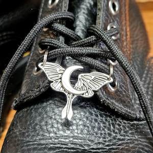 May include: Close-up of a black leather boot with black laces. A silver-colored charm in the shape of a moth with a crescent moon is attached to the laces. The boot has visible stitching and metal eyelets.
