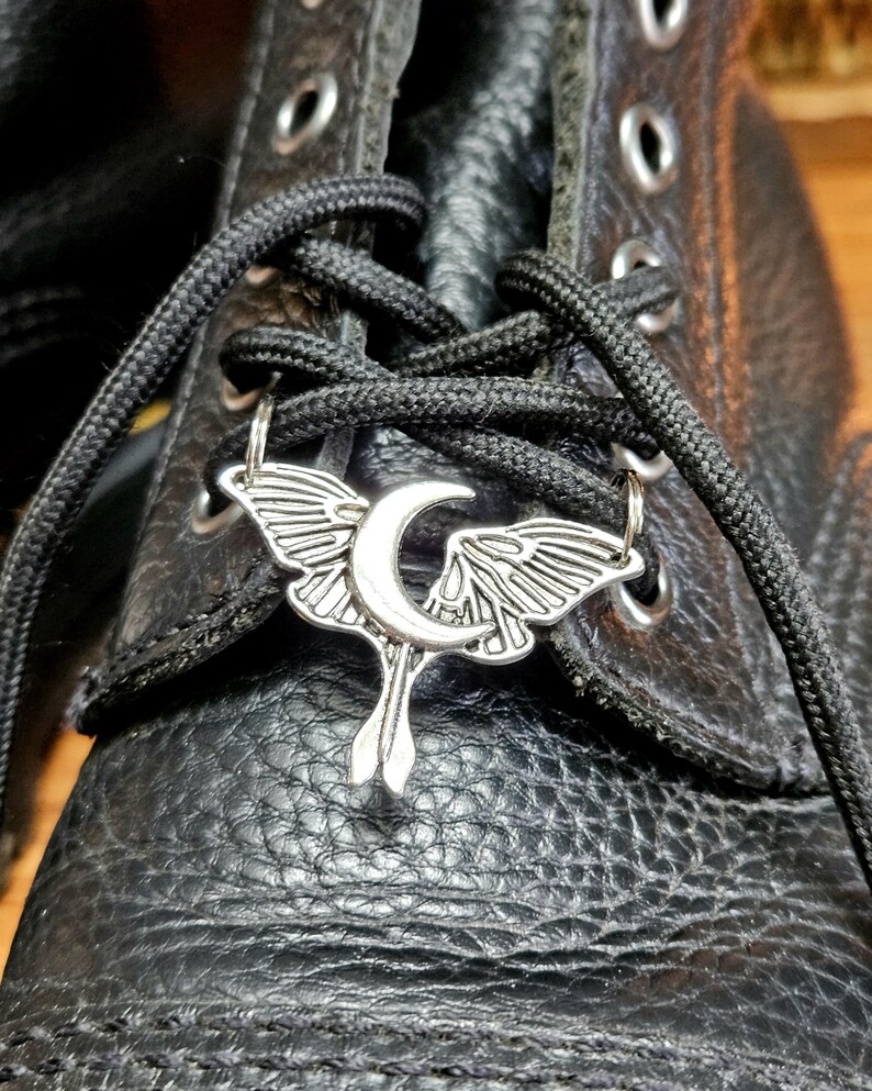 May include: Close-up of a black leather boot with black laces. A silver-colored charm in the shape of a moth with a crescent moon is attached to the laces. The boot has visible stitching and metal eyelets.