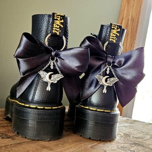 Könnte beinhalten: Schwarze Plateau-Stiefel mit gelben Nähten und einer schwarzen Satinschleife. Jeder Stiefel hat einen silbernen Anhänger mit Mond und Nachtfalter-Design. Der Text "AirWair" ist auf der Lasche zu sehen.