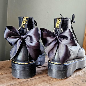 May include: Two black platform boots with black satin bows. The boots feature yellow stitching and the brand name "Dr. Martens" in yellow. The boots are displayed on a wooden surface.