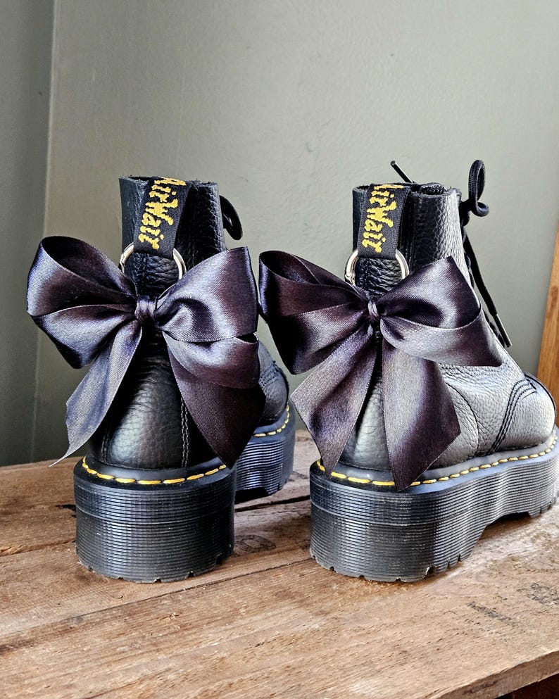 May include: Two black platform boots with black satin bows. The boots feature yellow stitching and the brand name "Dr. Martens" in yellow. The boots are displayed on a wooden surface.