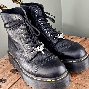 May include: Pair of black leather platform boots with silver-tone charms. The boots have a textured finish, black laces, and yellow stitching around the soles. The soles are thick and chunky.