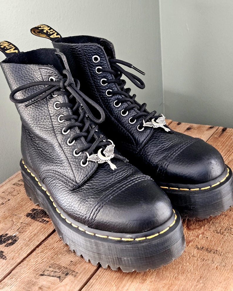 May include: Pair of black leather platform boots with silver-tone charms. The boots have a textured finish, black laces, and yellow stitching around the soles. The soles are thick and chunky.