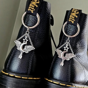 Może przedstawiać: Czarne skórzane buty z żółtymi przeszyciami i srebrnym urokiem. Urok przedstawia motyla z księżycem i gwiazdą, zawieszony na srebrnym pierścieniu przymocowanym do pętli buta.