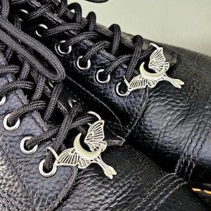 May include: Close-up of black leather boots with black laces and silver charms. The charms are shaped like a crescent moon with a butterfly. The boots have silver eyelets.