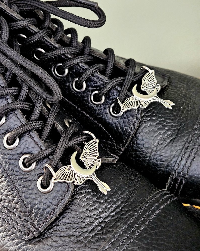 May include: Close-up of black leather boots with black laces and silver-colored charms. The charms are shaped like a crescent moon with a butterfly. The boots have silver eyelets.