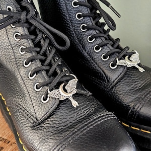 May include: Close-up of two black leather boots with black laces and silver moth-shaped charms. The boots have silver eyelets and yellow stitching along the soles. The leather has a textured appearance.