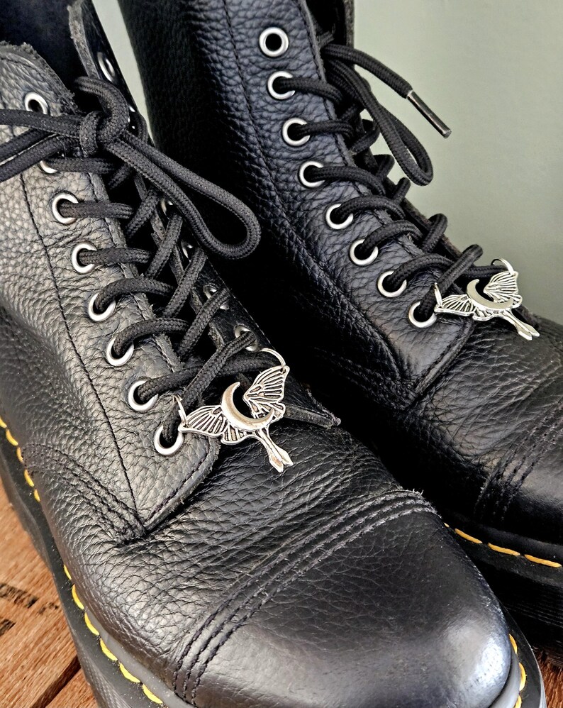 May include: Close-up of two black leather boots with black laces and silver moth-shaped charms. The boots have silver eyelets and yellow stitching along the soles. The leather has a textured appearance.
