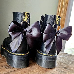 May include: Two black platform boots with black satin bows attached. The boots feature yellow stitching and the brand name is visible on the pull tab. The boots are displayed on a wooden surface.