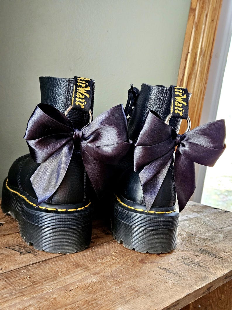 May include: Two black platform boots with black satin bows attached. The boots feature yellow stitching and the brand name is visible on the pull tab. The boots are displayed on a wooden surface.