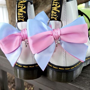 Puede incluir: Un par de botas de cuero blanco con tiradores negros y texto amarillo AirWair. Cada bota está adornada con un gran lazo de satén superpuesto en rosa pastel y azul claro, sujeto a una anilla de metal plateado con un pequeño dije de corazón.