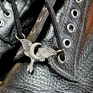 May include: Close-up of a black leather boot with a silver-colored charm. The charm features a crescent moon and stylized wings, attached to the black laces. The boot has multiple eyelets and a textured surface.