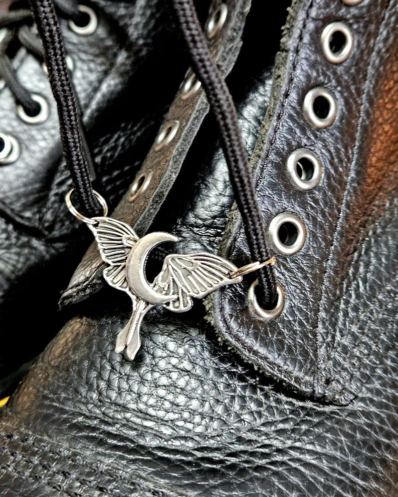 May include: Close-up of a black leather boot with a silver-colored charm. The charm features a crescent moon and stylized wings, attached to the black laces. The boot has multiple eyelets and a textured surface.