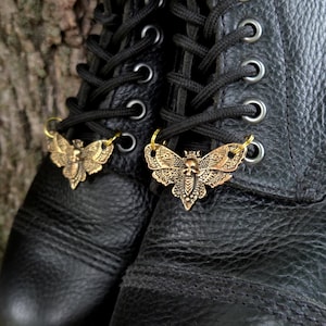 May include: A pair of black leather lace-up boots featuring two brass-toned metal moth-shaped charms attached to the laces. Each charm displays an intricate wing pattern with a central skull detail, secured by small gold-colored jump rings.