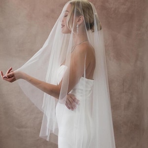 May include: A sheer, floor-length white bridal veil made of fine tulle fabric. The veil is draped over the head and shoulders, extending down the back. It is paired with a strapless white gown, a delicate silver necklace, and drop earrings.